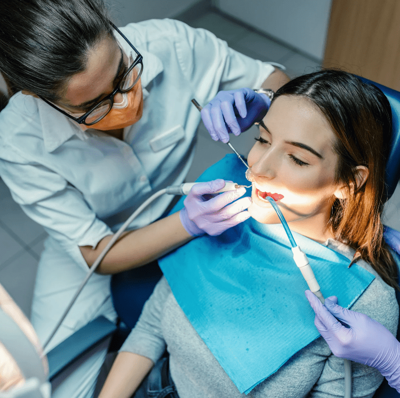 can-dentists-remove-tonsil-stones A female patient receiving dental treatment from a dentist wearing a mask and gloves. The dentist uses dental tools while an assistant holds a suction device.