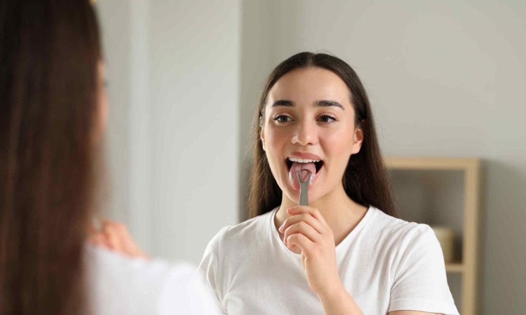 woman-using-tongue-scraper Comment utiliser un gratte-langue: une femme devant son miroir utilise un gratte-langue en ouvrant la bouche et en tirant la langue